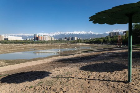 Empty City sand beach with drained pond. Metal stationary sun umbrellas. Residential apartment buildings and high snow-capped mountains in background. Storage reservoir Lake Sayran Almaty Kazakhstan. 