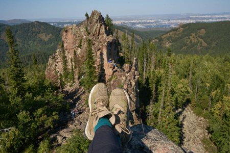 Viajero descansando en la cima de un acantilado formación de rocas ígneas. Paisaje natural de verano, Parque nacional de Stolby, Piernas humanas en botas de senderismo. Colinas en el bosque. Krasnoyarsk, Siberia, Rusia. Estilo de vida