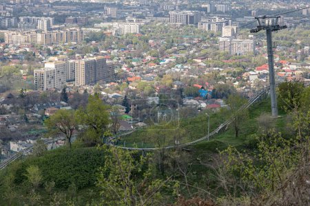 telesilla vacía que conduce al parque de atracciones Kok Tobe y rodelbahn en la ladera de la montaña. Hierba verde y arbustos, soleado día de primavera. Ver edificios Almaty ciudad Kazajstán Paisaje urbano. cable de tracción portante