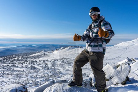 Heureux homme souriant dans l'équipement de ski d'hiver montre pouces levés, recommandation bon choix. Veste chaude, pantalon, gants marron, lunettes de ski, bottes, casque de sport. sur toute la longueur. Activités Sheregesh resort