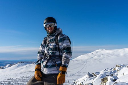 Satisfied man at Sheregesh ski resort, against backdrop snowy slopes of mountain at sunny day. Winter travel activities. Men in snowboard equipment black helmet, goggles, membrane jacket, brown gloves