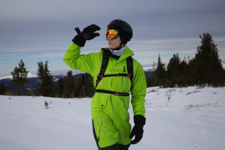 Man in bright acid green snowboard suit, black warm gloves, orange goggles, helmet and backpack skier outfit, reaches hand towards his had. Explores surroundings of ski resort. get lost pathfinding