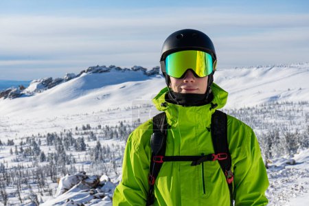 Close-up portrait of a skier or snowboarder in sports equipment, snowy mountains background at ski resort. Bright acid green outfit: warm suit jacket, goggles, black sport helmet, backpack straps