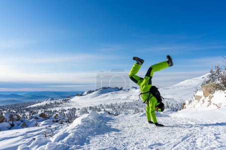 Man stands on his hands upside down Bright acid green outfit: warm suit, goggles, black helmet. Snowy mountains background at Sheregesh ski resort. Active lifestyle, winter leisure. Mountain View