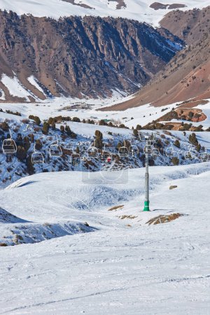 People sit in chairs of cable car leading to the top of ski slope. Ropeway pole, construction. Winter vacation activity, skiers, snowboarders, lifestyle. Resort Chunkurchak, Nature of Kyrgyzstan. 