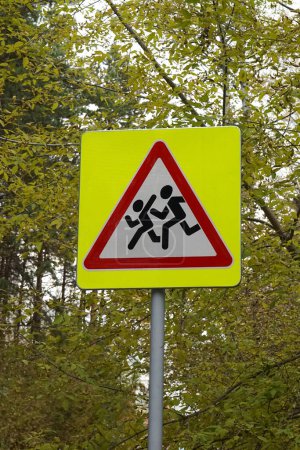 Road sign on pole, that is installed near schools. Denoting Be careful, children. Yellow square, red triangle, white, with the image of running people. Safety, Traffic warning, autumn forest.