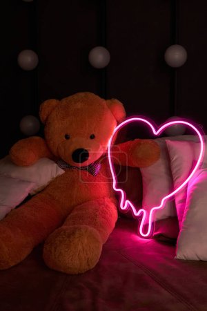 Brauner Teddybär liegt in Kissen und hält ein neonpinkfarbenes Herz. Valentinstag 14. Februar, Geschenk romantischen Hintergrund. Liebeserklärung, Glückwunsch zum Urlaub oder Jubiläum. 