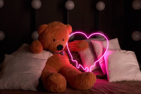 Brown teddy bear lies in pillows and holds a neon pink heart. Valentine's Day 14 February, Gift romantic background. Declaration of love, congratulations on the holiday or anniversary. 