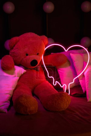 Niedlicher brauner Teddybär liegt in Kissen, hält ein leuchtendes neonpinkfarbenes Herz. Valentinstag 14. Februar, Geschenk romantischen Hintergrund. Liebeserklärung, Glückwunsch zum Urlaub oder Jubiläum