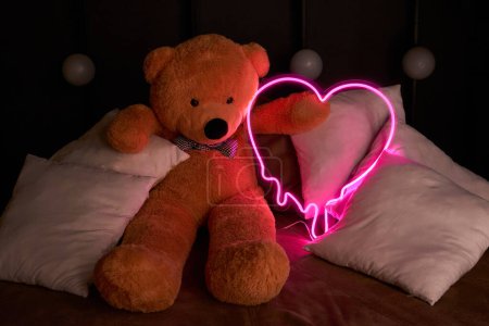 Brown teddy bear lies in pillows and holds a neon pink heart. Valentine's Day 14 February, Gift romantic background. Declaration of love, congratulations on the holiday or anniversary. 