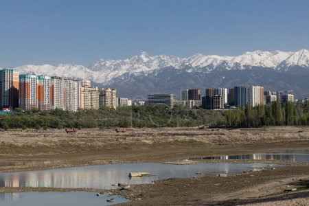 Storage reservoir Lake Sayran, Almaty, Kazakhstan. Empty City sand beach with drained pond. Residential apartment buildings and high snow-capped mountains in background. Recreation place for citizens