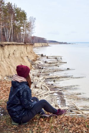 Woman in warm clothes, sits on cliff above coast, admires nature, enjoy solitude. Active Lifestyle moment. Concept of traveling in any weather, regardless of season, free spirit. Winter, autumn leaves