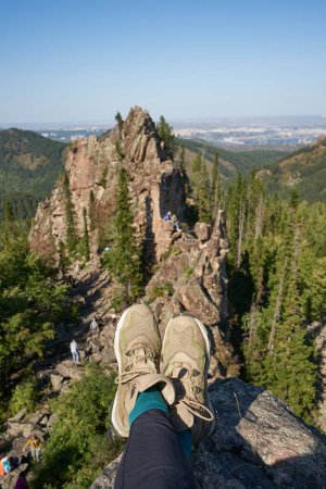 Reach the top of cliff. Legs of a man in hiking boots against the background of igneous rocks and green forest near the city. Danger of sitting on the edge of rock without insurance. Amazing nature