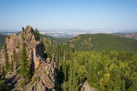 Eine Gruppe Touristen sitzt auf einem glühenden Felsen vor der Kulisse grüner Wälder und Städte in der Ferne. Sommerliche Naturlandschaft. Aktiver Lebensstil: Klettern, Wandern und Tourismus. Die Natur genießen
