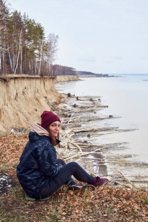 Junge Frau in warmen Kleidern, sitzt auf einer Klippe über der Küste, blickt in die Kamera, bewundert die Natur, genießt die Einsamkeit. Aktiver Lebensstil Reisemoment. Konzept des Reisens bei jedem Wetter, unabhängig von der Jahreszeit
