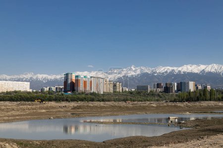 Urban landscape, modern buildings rise against the awe-inspiring backdrop of snow-capped mountains. Clear blue sky reflects off pond water. Unique beauty of a city nestled at foot of towering peaks