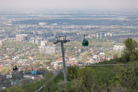 Cable car with two cabins, against the backdrop of the city at spring time. Aerial lift to Kok Tobe hill in Almaty, Kazakhstan. Tourist place, city landmark. Ropeway support pillar