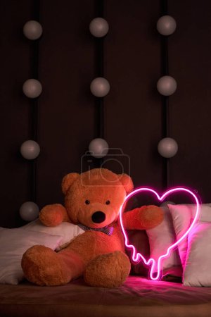Cute brown teddy bear lies in pillows, holds a brightly glowing neon pink heart. Valentines Day 14 February, Gift romantic background. Declaration of love, congratulations on holiday or anniversary