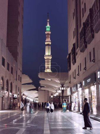 Photo for AL MADINAH, SAUDI ARABIA-MAY 24, 2023 : Low light evening view one of the Nabawi Mosque minarets with Muslim pilgrims walking at the entrance of Nabawi mosque in Medina, Saudi Arabia. - Royalty Free Image