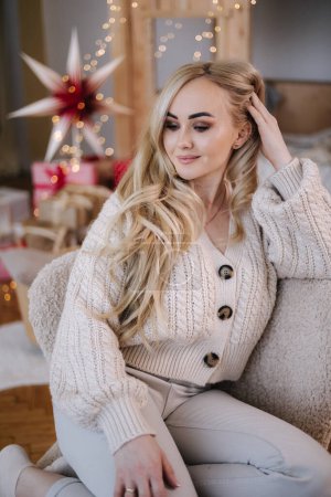 Téléchargez les photos : Femme aux cheveux blonds en costume tricoté blanc assis à la maison près du sapin de Noël. Belle femme en vacances, humeur de Noël. - en image libre de droit