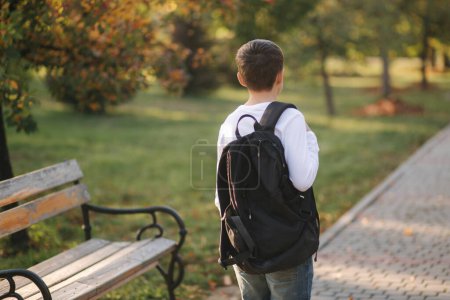 Back view of teenage boy go to school. Young male in white sweatshirt with backpack go though the park.