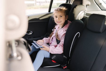 Photo for Adorable child girl playing game with digital tablet while sitting in car seat of car during trip. Happy female kid enjoys vacation with tablet in car. High quality photo - Royalty Free Image