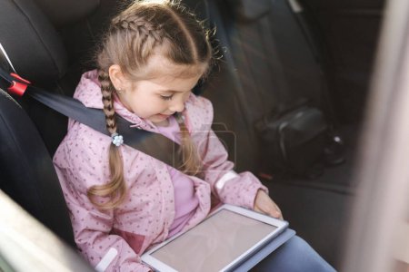 Photo for Beautiful little girl in car seat using digital tablet during trip. Happy female kid on back seat. High quality photo - Royalty Free Image