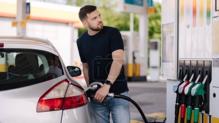 Foto de Hombre barbudo repostando coche en la gasolinera y mirando en su teléfono inteligente. El hombre compara los precios del combustible. Foto de alta calidad - Imagen libre de derechos