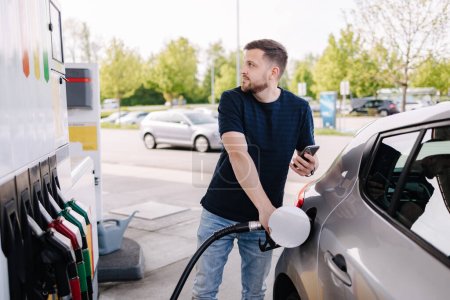 Foto de Hombre barbudo guapo repostando coche y mirando en el marcador mientras está de pie en la gasolinera de autoservicio. Foto de alta calidad - Imagen libre de derechos