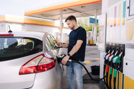 Foto de Hombre barbudo repostando coche en la gasolinera y mirando en su teléfono inteligente. El hombre compara los precios del combustible. Foto de alta calidad - Imagen libre de derechos