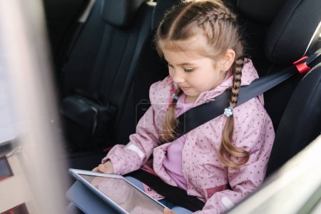 Photo for Cute little girl use tablet in car seat on the way to the kindergarten. High quality photo - Royalty Free Image