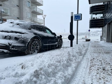 Foto de Estocolmo, Suecia - 8 de marzo de 2023: Coche eléctrico cargando en la calle fuera del edificio residencial en el clima de nieve en la ciudad. Nieva en el día de invierno con el coche estacionado en la nieve. - Imagen libre de derechos