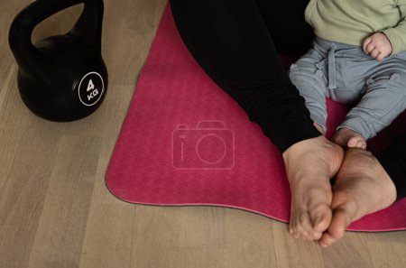 Foto de Bebé y mujer sentados en una esterilla de yoga listos para el ejercicio posparto con pesas de 4 kg. Concepto de entrenamiento con bebé o niño pequeño. Estilo de vida activo con entrenamiento de fuerza después del parto. - Imagen libre de derechos