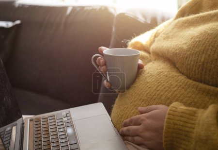Foto de Mujer embarazada sentada en el sofá acariciando su vientre con un portátil en su regazo y sosteniendo una taza de café. Concepto de mujer embarazada trabajando desde casa por la mañana con la luz del sol. Foto tomada en Suecia. - Imagen libre de derechos