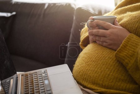 Foto de Mujer embarazada sentada en el sofá con portátil en su regazo y sosteniendo taza de café. Concepto de mujer embarazada trabajando desde casa por la mañana con la luz del sol. Foto tomada en Suecia. - Imagen libre de derechos