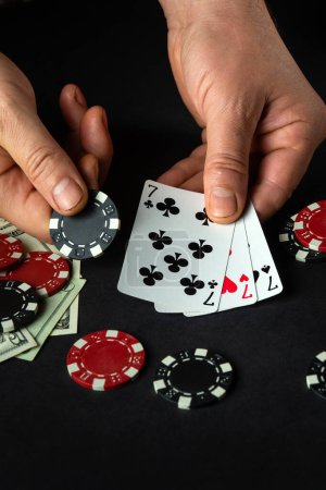 Foto de El jugador pone cartas de juego con una combinación ganadora de tres de una clase o se establece en una partida de póquer en una mesa negra con fichas y dinero en un club. - Imagen libre de derechos