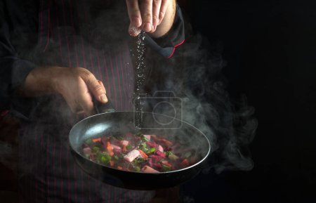 Photo for The chef salts vegetables in a hot frying pan. Menu concept for hotel or restaurant on black background - Royalty Free Image