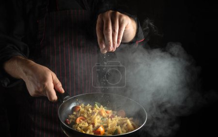 Photo for The chef adds salt to a steaming hot pan with vegetables and pasta. Thai restaurant cuisine or street food - Royalty Free Image