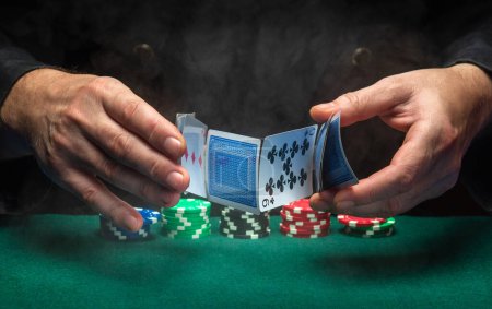 Foto de Close-up of the hands of a dealer or croupier shuffling poker cards in a smoky club against a green table with chips. Concept of playing poker or gaming business. - Imagen libre de derechos