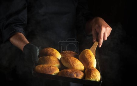 Foto de Professional baker is baking pies or hot dog buns on a hot baking sheet. Black space for menu or recipe - Imagen libre de derechos
