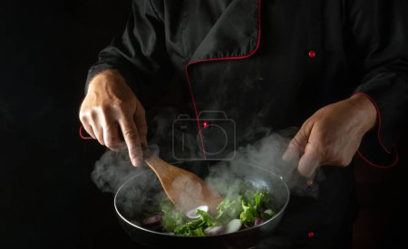 Photo for A professional chef prepares vegetables in a frying pan. The concept of cooking healthy vegetarian food and meals on a dark background. Free advertising space. - Royalty Free Image