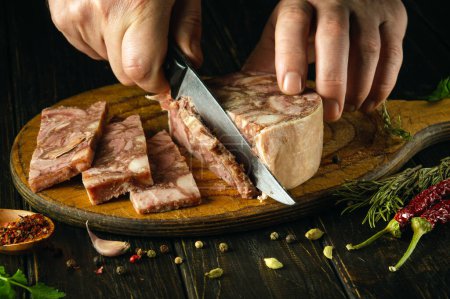 Photo for The cook cuts the brawn with a knife on the kitchen board before preparing sandwiches. Idea for a delicious snack with headcheese and aromatic spices - Royalty Free Image