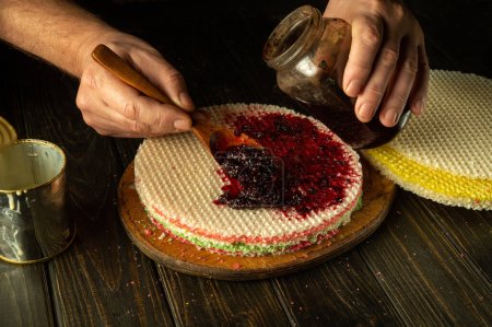 The chef is preparing a round waffle cake on the kitchen table. Adding jam with a spoon in the cook hand.