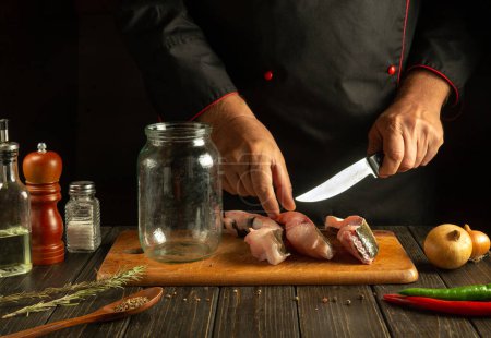 Photo for Slicing carp fish with a knife in the chef hand. The process of preparing a fish dish or herring by the hands of a cook on the kitchen table. - Royalty Free Image