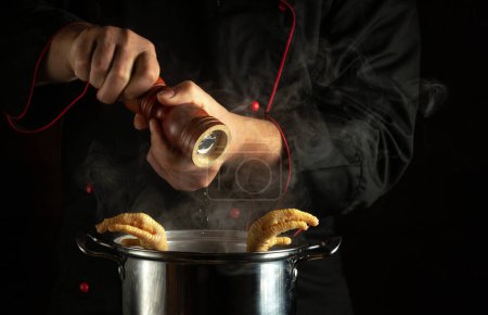 Téléchargez les photos : Un cuisinier en uniforme noir ajoute du poivre à une casserole de cuisses de poulet bouillantes. Le concept de préparer une soupe délicieuse et riche dans la cuisine. - en image libre de droit