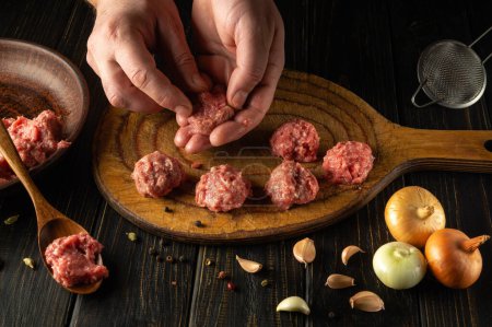 Chef shapes ground meat to make homemade meatballs for lunch. Concept of cooking food on kitchen table with meat vegetables and spices.