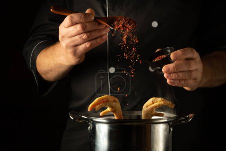 Téléchargez les photos : Le chef ajoute des épices rouges sèches dans une casserole de pieds de poulet mijotants. Restaurant cuisine concept de cuisine avec espace publicitaire sur fond noir. - en image libre de droit