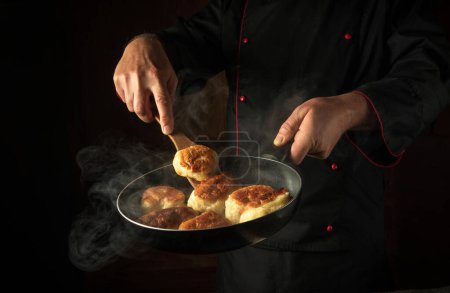 Ein Koch frittiert in einer Restaurantküche Donuts. Ein Küchenspachtel in der Hand eines Kochs und eine Pfanne mit Dampf. Konzept auf schwarzem Hintergrund des Kochens leckerer Pfannkuchen.