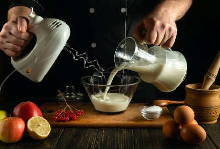 A professional chef prepares a milkshake with fruits in the kitchen using a hand-held electric mixer. The cook hand pours milk into a deep plate.