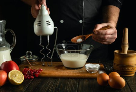 Milch und Zucker mit dem Handrührgerät schlagen. Low-Key-Konzept des Prozesses der Zubereitung eines Milchgerichts auf einem Küchentisch in einem öffentlichen Haus.